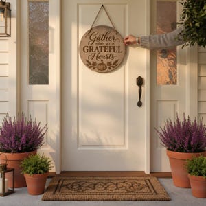 Può includere: Una targa da parete rotonda in legno appesa a una porta bianca con una corda. La targa presenta la scritta "Gather HERE WITH GRATEFUL HEARTS" in lettere marrone scuro, accompagnata da un'illustrazione di una zucca e foglie.