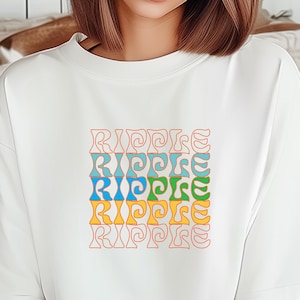 May include: White t-shirt with the word "RIPPLE" in a colorful, retro font. The word is stacked vertically in a square, with each instance of the word in a different color: pink, blue, teal, green, yellow, and orange.