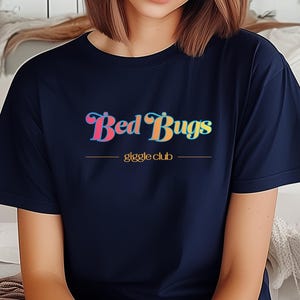 May include: Navy blue t-shirt with the text "Bed Bugs" in a colorful, retro font above the words "giggle club" in a smaller font. The shirt is a solid color and has short sleeves.