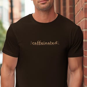 Pode incluir: Camiseta marrom escuro com a palavra "caffeinated" em marrom claro, com um estilo de escrita à mão. A camiseta de manga curta parece ser feita de um material macio.