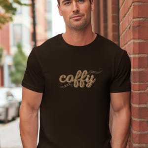 Könnte beinhalten: Ein dunkelbraunes T-Shirt mit dem Wort "coffy" in einer stilisierten, kursiven Schrift. Der Text ist hellbraun. Das kurzärmelige Shirt scheint aus einem weichen Material zu sein.