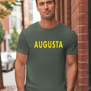May include: A short-sleeved, olive green t-shirt with the word "AUGUSTA" printed in yellow block letters. The shirt is made of a soft material and is being worn by a person. The background includes a brick wall and a blurred outdoor scene.