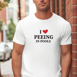 Puede incluir: Camiseta blanca con el texto "I ❤️ PEEING IN POOLS" en negro y un corazón rojo. La camiseta es de cuello redondo. La imagen muestra la camiseta en una persona. La tela es probablemente de algodón.