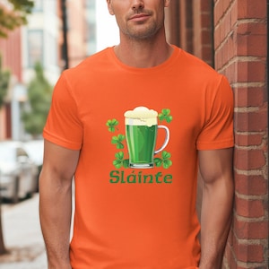 以下が含まれることがあります： オレンジ色のTシャツ。緑色のビールジョッキのデザインで、白い泡と緑のクローバーが描かれています。「Sláinte」の文字が緑色でプリントされています。Tシャツは無地のオレンジ色です。