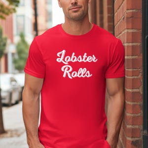 Może przedstawiać: Czerwona koszulka z napisem "Lobster Rolls" w białym skrypcie. Koszulka z krótkim rękawem wydaje się być wykonana z miękkiego materiału. Tekst jest wyśrodkowany na klatce piersiowej.