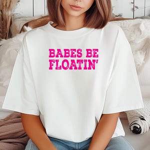 Puede incluir: Una camiseta blanca con la frase "BABES BE FLOATIN'" en rosa brillante en el centro. La camiseta tiene un corte holgado y mangas cortas. El estilo es casual y cómodo.