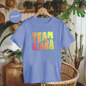 Puede incluir: Una camiseta violeta con el texto "TEAM CIARA" en una fuente colorida y de burbujas, delineada en azul y con un gráfico floral. La camiseta se muestra en una percha de madera.
