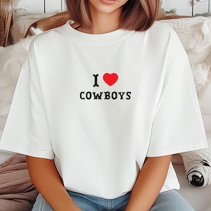 May include: White t-shirt featuring the text "I ♥ COWBOYS" in black and red. The heart symbol is red. The shirt is a casual style, suitable for everyday wear.