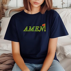 Puede incluir: Camiseta azul marino con la palabra "AMEN" en verde y amarillo, acompañada de un pequeño gráfico de bandera de golf. La camiseta es de estilo casual, adecuada para el uso diario. El texto está en una fuente estilizada.