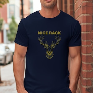 Puede incluir: Camiseta azul marino con un gráfico de cabeza de ciervo dorado y el texto "NICE RACK". El gráfico presenta una ilustración detallada de los cuernos y la cara de un ciervo. La camiseta está hecha de un material suave y cómodo.