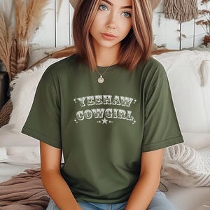 May include: An olive green t-shirt with the text "YEEHAW COWGIRL" in a decorative font. The text is embellished with stars. The shirt is a casual style.