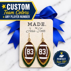May include: A pair of football-shaped dangle earrings featuring a brown textured surface with white laces and the number 83 printed in white. The earrings are layered over a gold glitter backing and hang from gold-toned metal hooks on a display card.
