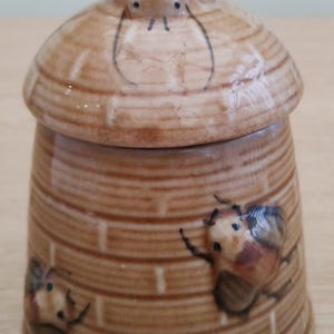 May include: A ceramic honey pot with a lid shaped like a beehive. The pot is brown with a textured, ribbed design. Painted bees decorate the sides. The lid has a small, pointed top.
