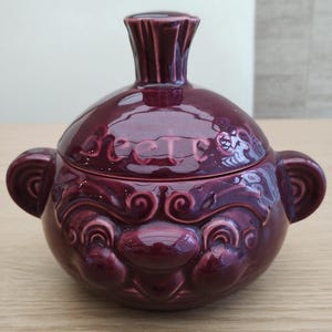 May include: A burgundy ceramic lidded bowl with a face design. The bowl has a bulbous shape with two ear-like handles and a tall, fluted neck. The face features a smiling expression with detailed, swirling patterns.