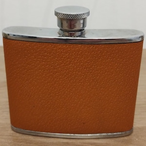 May include: A hip flask with a textured, orange-brown leatherette finish. It features a silver-coloured top and a screw-on cap. The flask is designed for carrying liquids and is a portable container, often used for spirits.