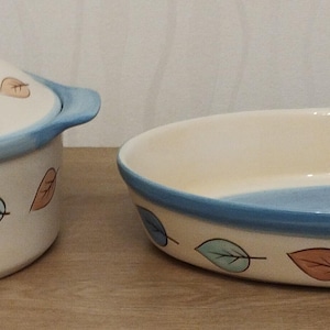 May include: A cream-coloured ceramic casserole dish with a lid and an oval baking dish. Both pieces have a blue rim and are decorated with leaf designs in orange, blue, and green. The casserole dish has a blue handle on each side.