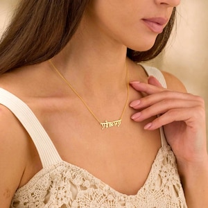 May include: A gold-colored necklace with a delicate chain. The pendant features an inscription in an elegant script. The necklace is worn around the neck.