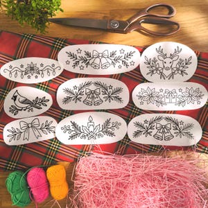Könnte beinhalten: Eine Auswahl weißer, ovaler Aufkleber mit schwarzen Strichzeichnungen von festlichen Motiven wie Glocken und Schleifen. Die Aufkleber liegen auf einer rot-grünen Tartan-Oberfläche, mit einer Schere und Garn im Hintergrund.