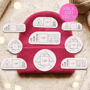 May include: A magenta beanie with several white stickers featuring camping-themed illustrations. The stickers include images of tents, mountains, campfires, and RVs, with measurements in inches. The text "STICK AND STITCH" is on a pink circle.