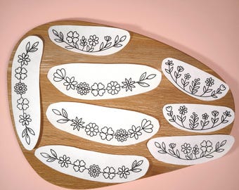 Könnte beinhalten: Eine Sammlung weißer Aufkleber mit Blumenmustern, angeordnet auf einem ovalen Holzbrett. Jeder Aufkleber zeigt eine andere Anordnung von schwarz gezeichneten Blumen und Blättern. Der Hintergrund ist einfarbig rosa.