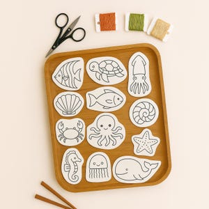May include: A wooden tray displays white, die-cut sea creature shapes such as a fish, turtle, and octopus. Above the tray are three spools of thread in orange, green, and tan. Black scissors and two wooden sticks are also present.
