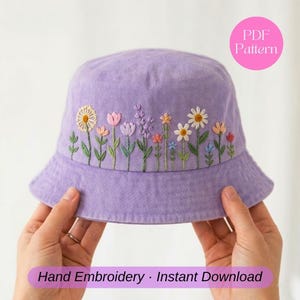 Puede incluir: Un sombrero de pescador lavanda con diseños florales bordados a mano. El sombrero presenta una variedad de flores coloridas, incluyendo margaritas, tulipanes y lavanda. El texto "PDF Pattern" está en un círculo rosa, y el texto "Hand Embroidery • Instant Download" está en una pancarta morada.