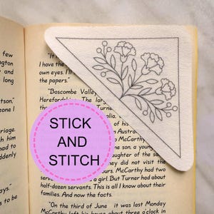 Puede incluir: Un marcapáginas triangular de tela blanquecina con un diseño de bordado floral en hilo gris. El marcapáginas está colocado en la esquina de un libro con texto. Las palabras "STICK AND STITCH" están en un círculo rosa.