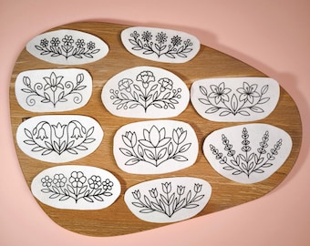 Könnte beinhalten: Eine Sammlung von neun weißen Stoffflicken mit schwarzen floralen Designs, angeordnet auf einem ovalen Holzbrett. Die floralen Muster umfassen verschiedene Blumensorten und Blattdetails, vor einem zartrosa Hintergrund.