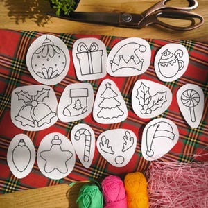 Puede incluir: Una colección de recortes navideños de papel blanco con contornos negros, que incluyen adornos, un regalo, un calcetín, un árbol y un bastón de caramelo. Los recortes están dispuestos sobre una tela a cuadros roja y verde, con tijeras e hilo en el fondo.