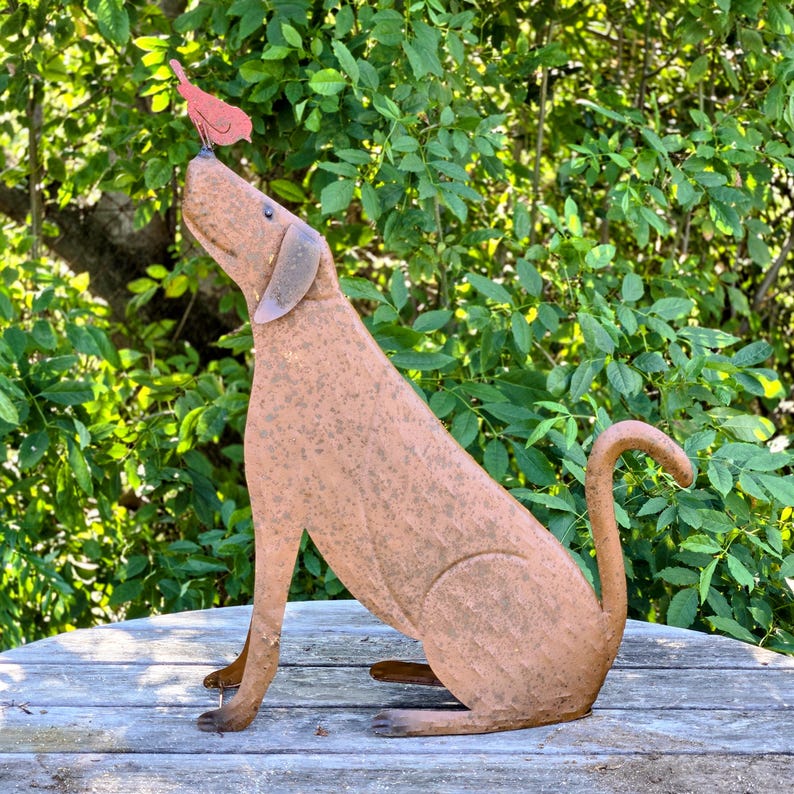 Peut inclure: Une sculpture de chien en m&eacute;tal, en position assise, avec un petit oiseau rouge perch&eacute; sur son nez. Le chien est de couleur marron rouille, avec une longue queue et des oreilles tombantes. La sculpture est &agrave; l'ext&eacute;rieur, avec un feuillage vert en arri&egrave;re-plan.