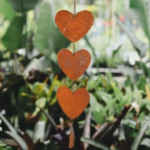 May include: A decorative hanging ornament with three heart-shaped cutouts made of rusty metal, linked by chains. The hearts hang against a backdrop of green foliage, creating a rustic and charming aesthetic.
