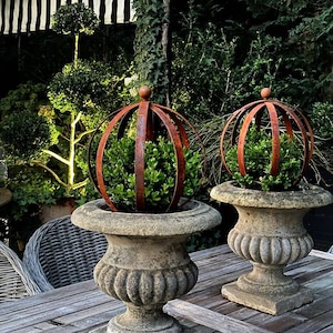 May include: Two weathered stone urns with green plants, topped with rusty metal orb-shaped cages. The urns sit on a wooden table, with a striped awning and lush greenery in the background. A decorative garden accent.