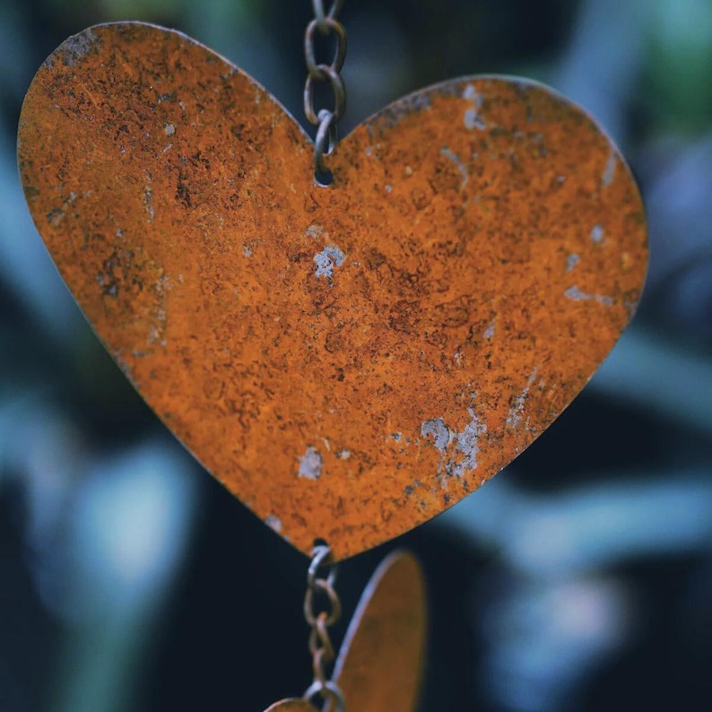 May include: Close-up of a heart-shaped metal ornament with a rusty orange finish. The heart is suspended by a chain, with a blurred background of green and blue foliage. The ornament has a textured surface.