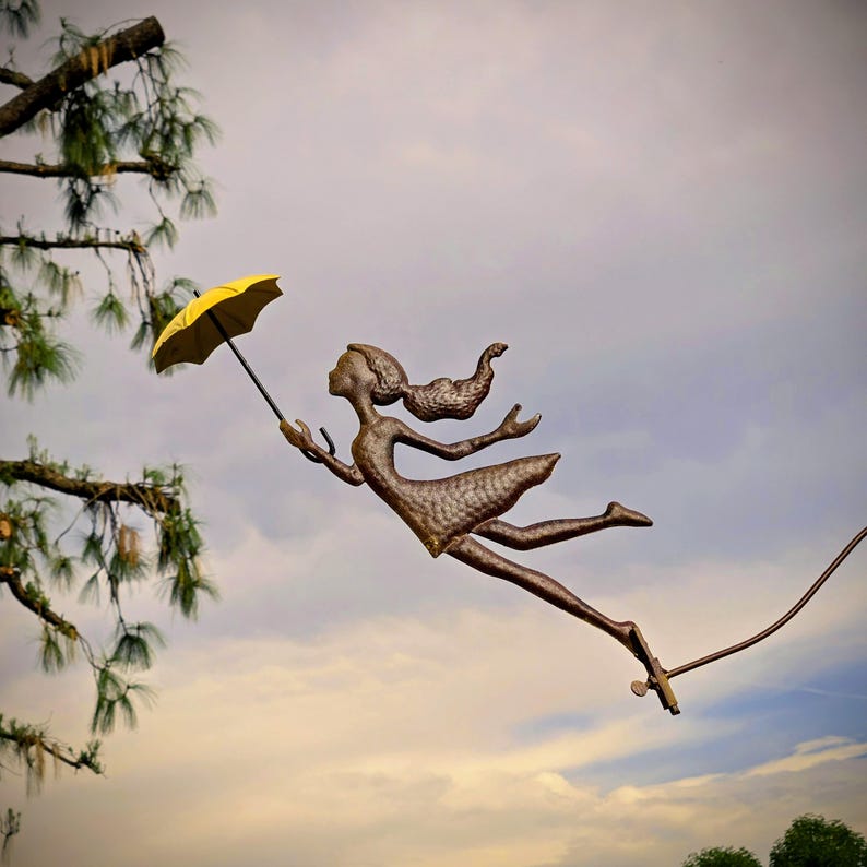 May include: A bronze sculpture of a figure holding a yellow umbrella, appearing to float in the air. The sculpture is set against a cloudy sky, with tree branches in the background. The figure is wearing a dress and has flowing hair.