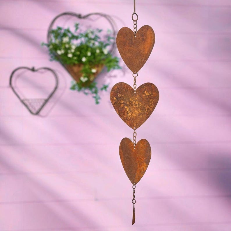 May include: A decorative hanging wind chime featuring three heart-shaped metal elements with a rusty brown finish, connected by chains. Two heart-shaped wire wall planters are also visible, one filled with greenery.
