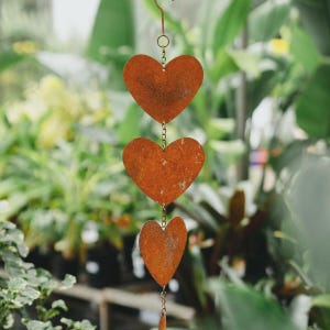 May include: A decorative hanging ornament with three heart-shaped metal pieces. The hearts are linked by chains and have a rusty, orange-brown colour. The background is blurred, showing green foliage.
