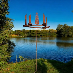 May include: A weathered metal sculpture of a lighthouse and sailboats stands tall against a serene lake and blue sky. The sculpture's rusty brown color contrasts with the vibrant green foliage and water, creating a striking outdoor art piece.
