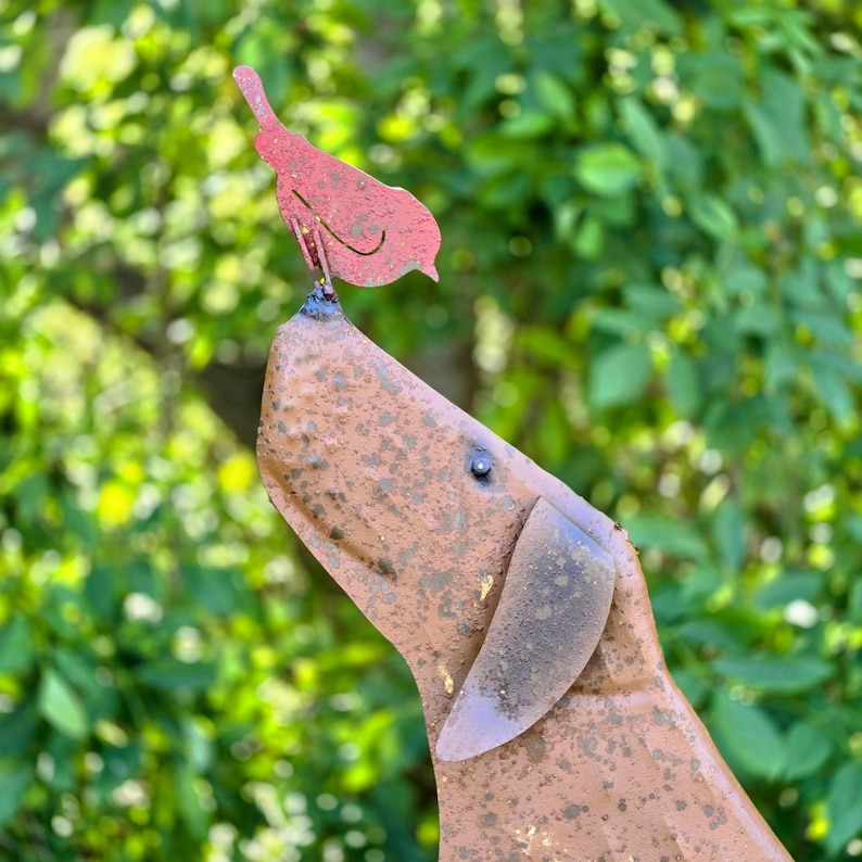 Peut inclure: Une sculpture m&eacute;tallique d'un chien regardant vers le haut, avec un oiseau rouge perch&eacute; sur son nez. La sculpture du chien a une finition marron vieillie et l'oiseau est de couleur rouge uni. Le fond est un feuillage vert flou.