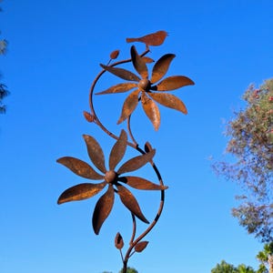 May include: A rustic metal garden sculpture featuring two large flower blooms and a small bird perched on top. The sculpture has a weathered, brown finish and is set against a clear blue sky. The flowers have multiple petals and are connected by a curved vine.