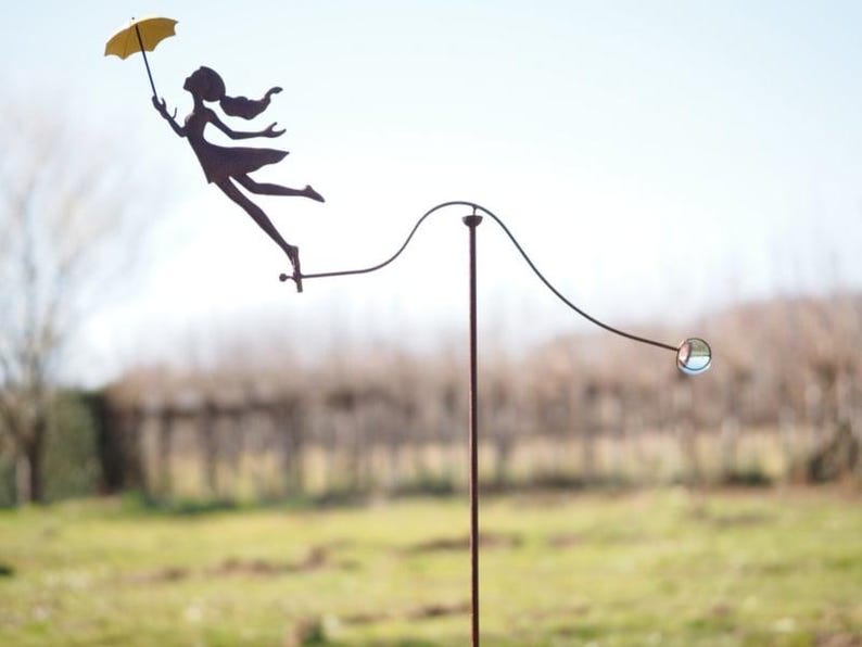 May include: A whimsical metal garden sculpture featuring a silhouette of a woman holding a yellow umbrella, seemingly floating in the air. The sculpture includes a curved rod with a glass ball at the end, set against a blurred outdoor background.