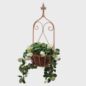 May include: A rustic, hanging planter with a brown, wrought-iron frame. The planter holds green and white flowering plants. The frame has a decorative top and scrollwork. The plants cascade down the sides of the planter.