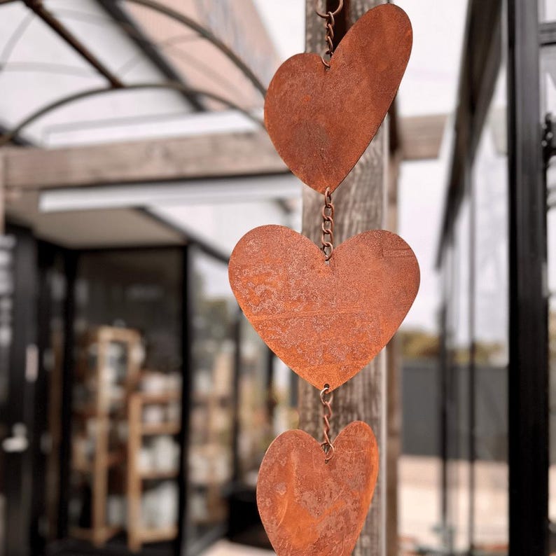 May include: Three rusty metal heart decorations suspended by a chain. The hearts vary in size and shape, with a weathered texture. The background is blurred, showing a building with glass windows.