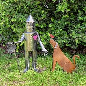 Peut inclure: Une sculpture m&eacute;tallique de l'Homme de fer-blanc du Magicien d'Oz, tenant une hache, avec un c&oelig;ur rose sur la poitrine. Une sculpture de chien en m&eacute;tal regarde un oiseau rouge. Les sculptures sont dans un jardin.