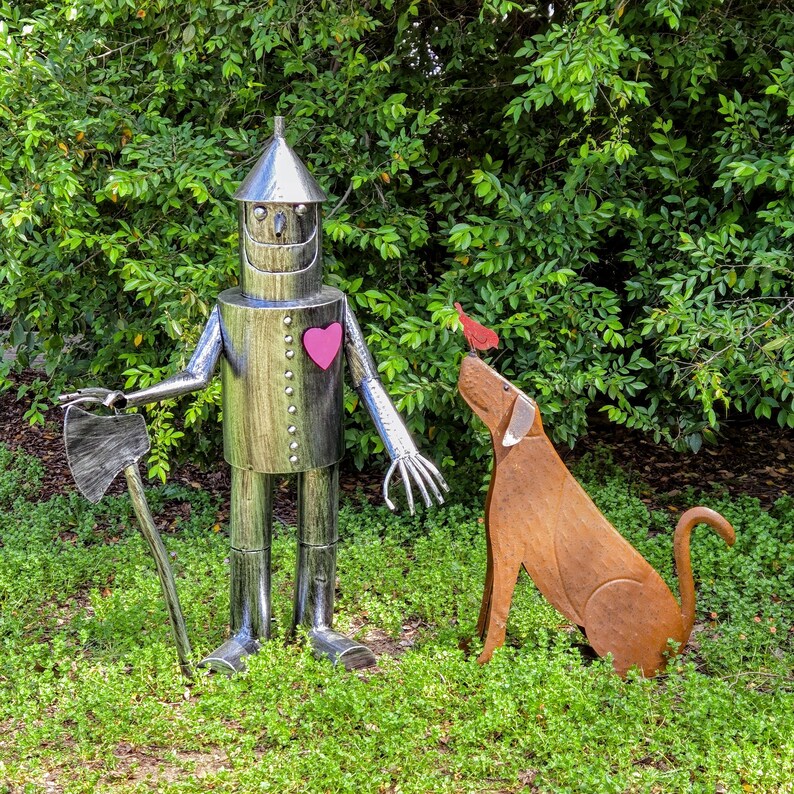 Peut inclure: Une sculpture m&eacute;tallique de l'Homme de fer-blanc du Magicien d'Oz, tenant une hache, avec un c&oelig;ur rose sur la poitrine. Une sculpture de chien en m&eacute;tal regarde un oiseau rouge. Les sculptures sont dans un jardin.