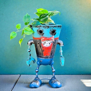 May include: A whimsical, robot-shaped planter with a distressed blue finish. The planter features a red bow tie, a black and red vest, and metal arms and legs. Green plants sprout from the top, adding a touch of life to the unique decor piece.