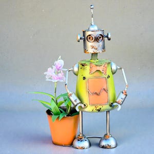 May include: A whimsical metal robot sculpture with a distressed, vintage appearance. The robot is painted in shades of green, orange, and silver, and features a bow tie and a small orange pot with a pink flower. The robot is holding the pot.