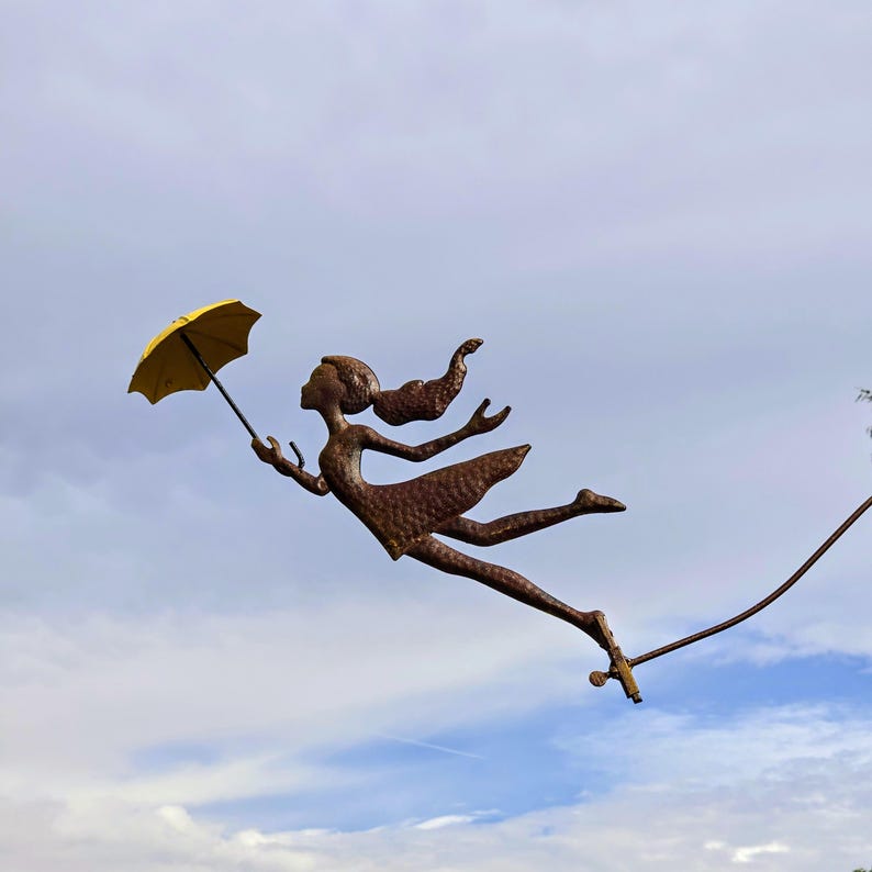 May include: A metal sculpture of a figure with flowing hair and a dress, holding a yellow umbrella, appears to be floating in the sky. The sculpture is a bronze color, and the background is a cloudy blue sky.
