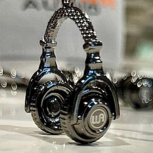May include: A silver-colored pair of headphones with a detailed design. The headphones have a black headband and earcups with a circular design. The earcups have a black and silver logo that says "WA".