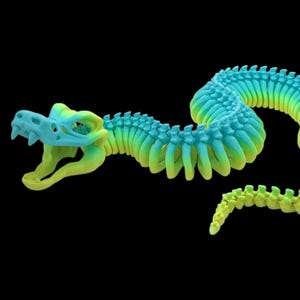 May include: A 3D-printed articulated snake toy in shades of blue and yellow-green. The snake's head is a light blue skull shape with open jaws. The body transitions from blue to yellow-green, with a segmented design. The background is black.