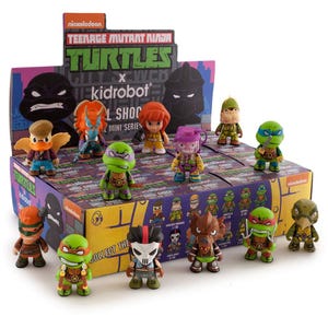 May include: A display box featuring Teenage Mutant Ninja Turtles x Kidrobot mini figures. The figures are colorful, with various characters from the series. The box has the Nickelodeon and Turtles logos, and the text "Shell Shock" and "Collect the".